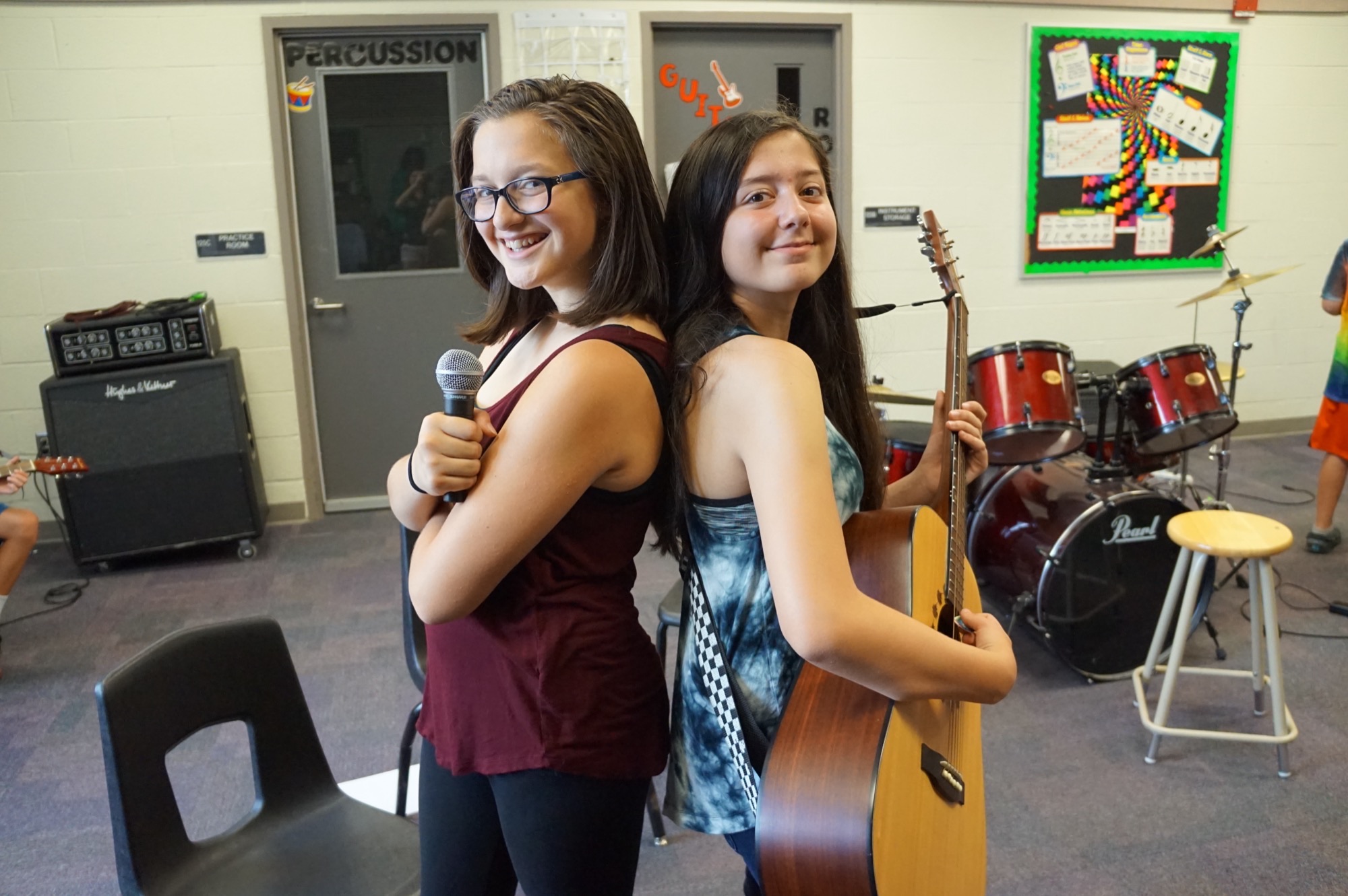 Two teenage rock-camp musicians stand back-to-back in a music room with a mic and an acoustic guitar — drumkit and amp visible behind them