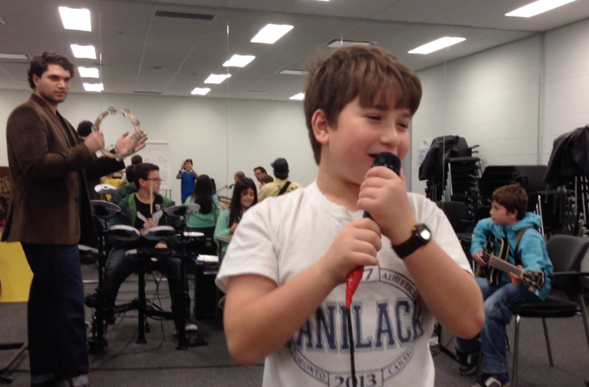 A young singer at the mic during a community-centre Rockstar session, with a teacher clapping and bandmates in the background