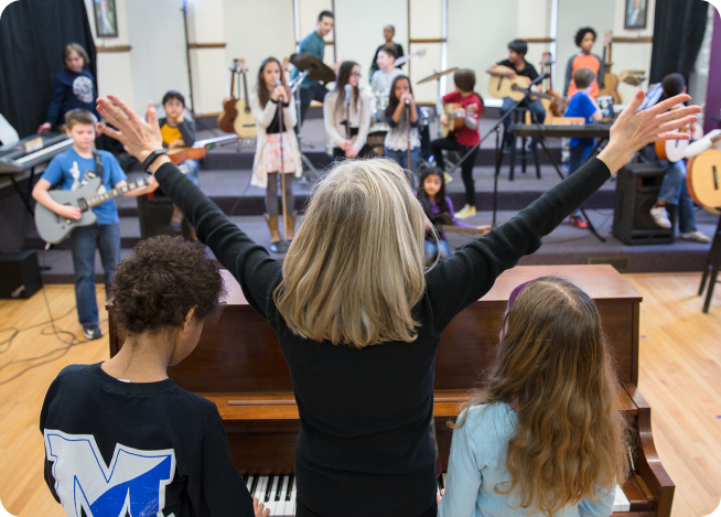 A Rockstar music director leads a full student band — kids on guitars, vocals, and piano — in a community music room