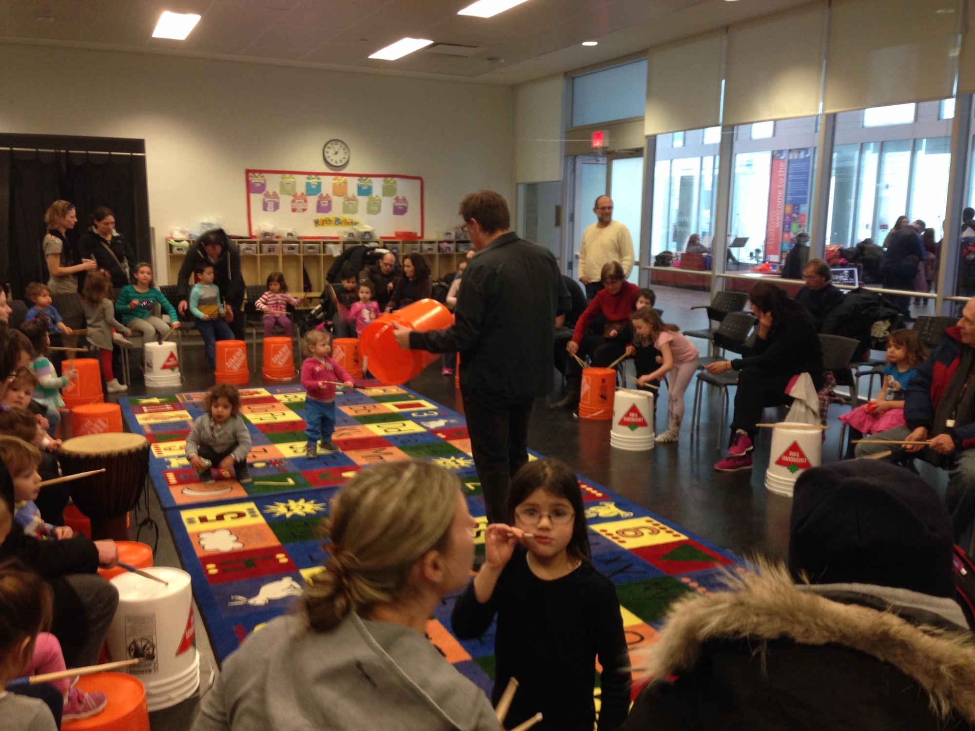 A Feel-the-Beat bucket-drumming workshop in full swing at a community library — dozens of children on Home Depot buckets, families watching, a Rockstar teacher leading from the centre