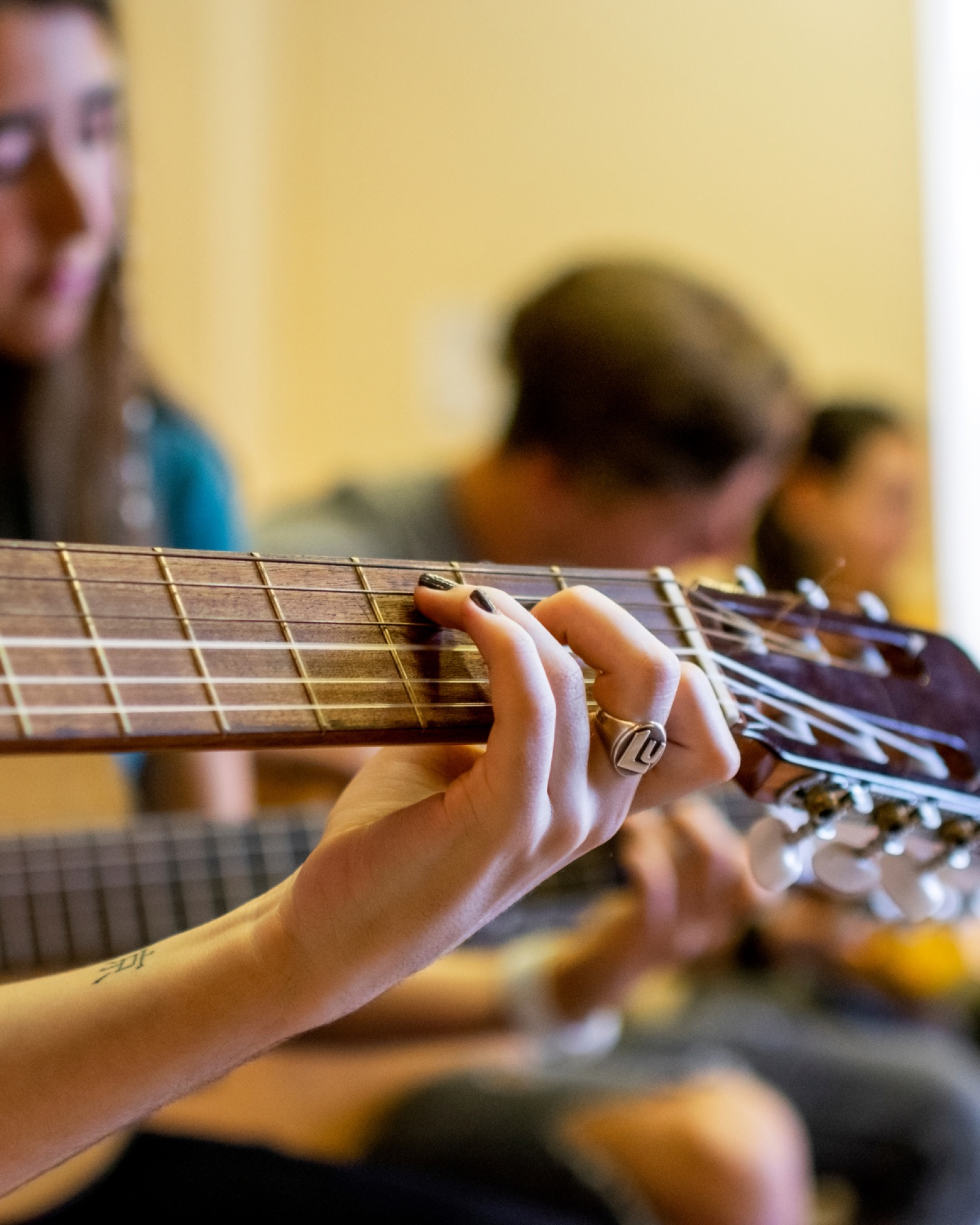 A student's hands on a classical guitar during a school music class — close-up on the fretboard with classmates playing in the background