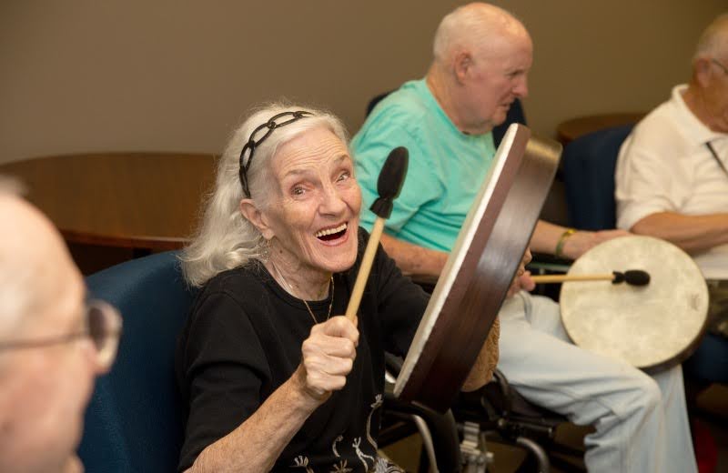 A senior beaming with joy mid-drum-strike during a Joy of Music session — fellow residents drumming alongside her in the residence common room